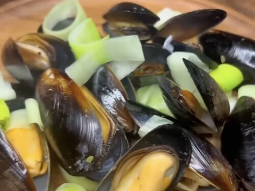 Cooked mussels with pale flesh visible, garnished with sliced leek and yellow vegetable pieces